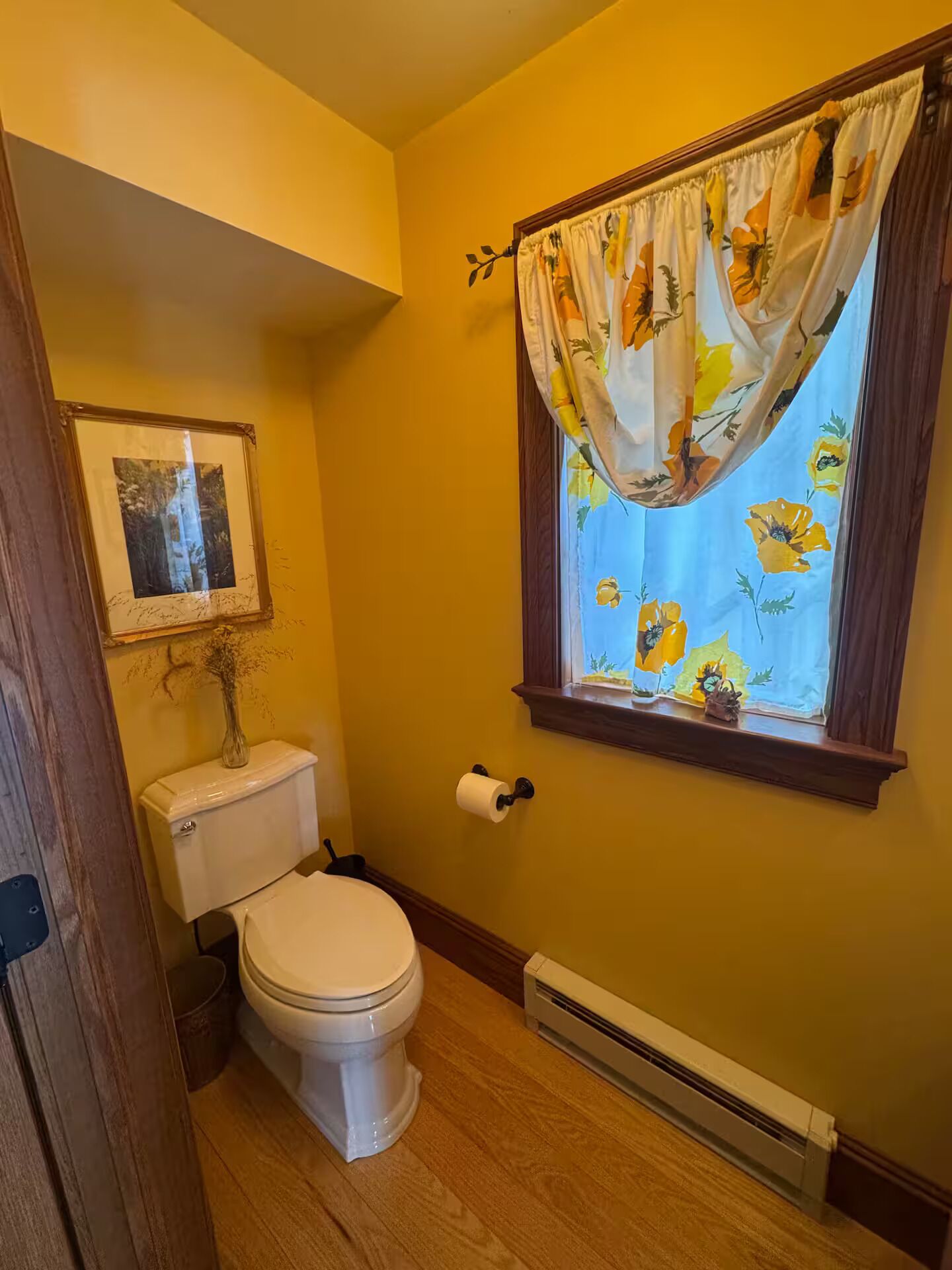 A small bathroom with a toilet, a framed picture on the wall, a window with floral curtains, and a radiator.