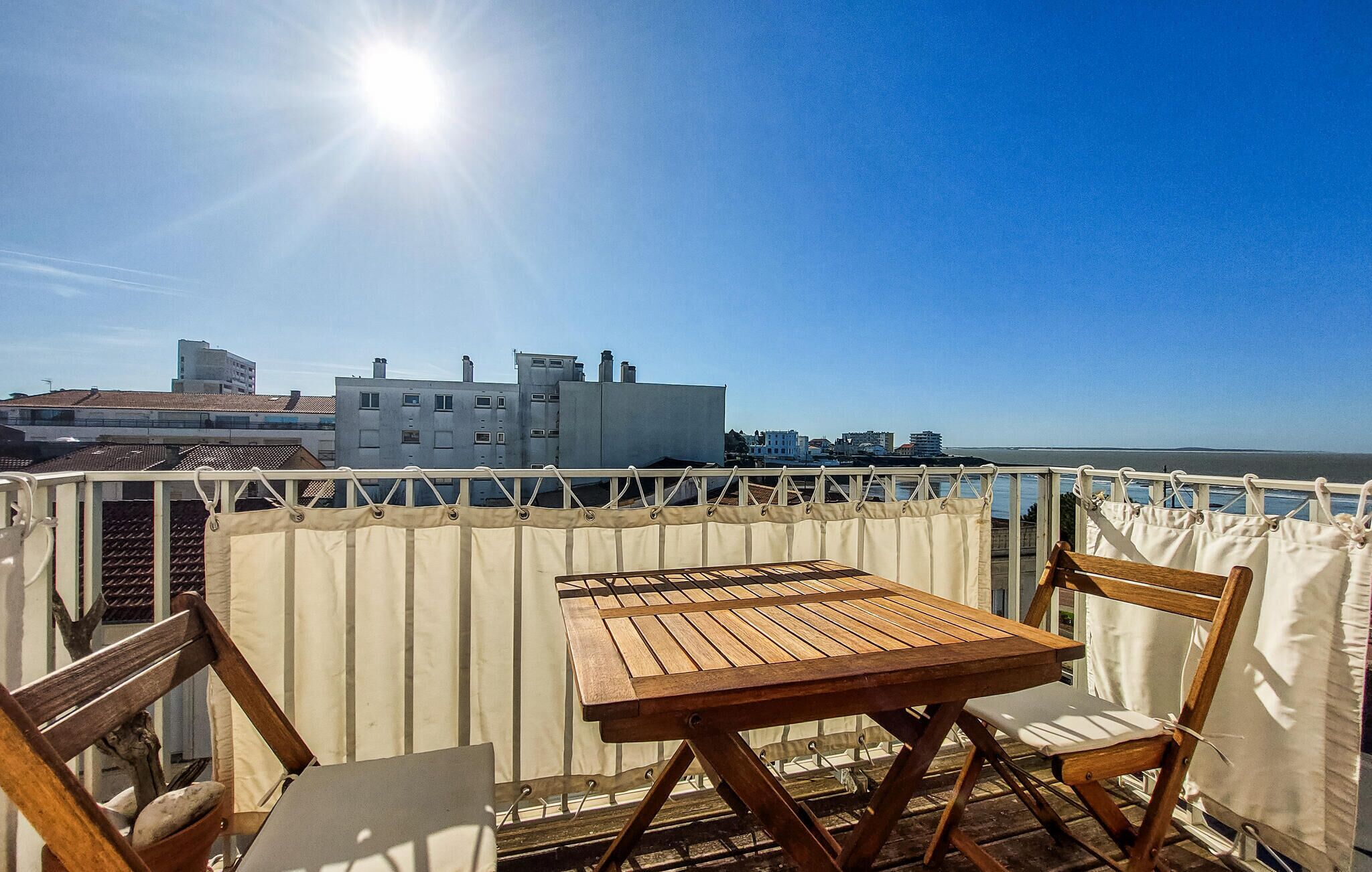 A wooden table and chairs on a balcony with a view of buildings and the sea.