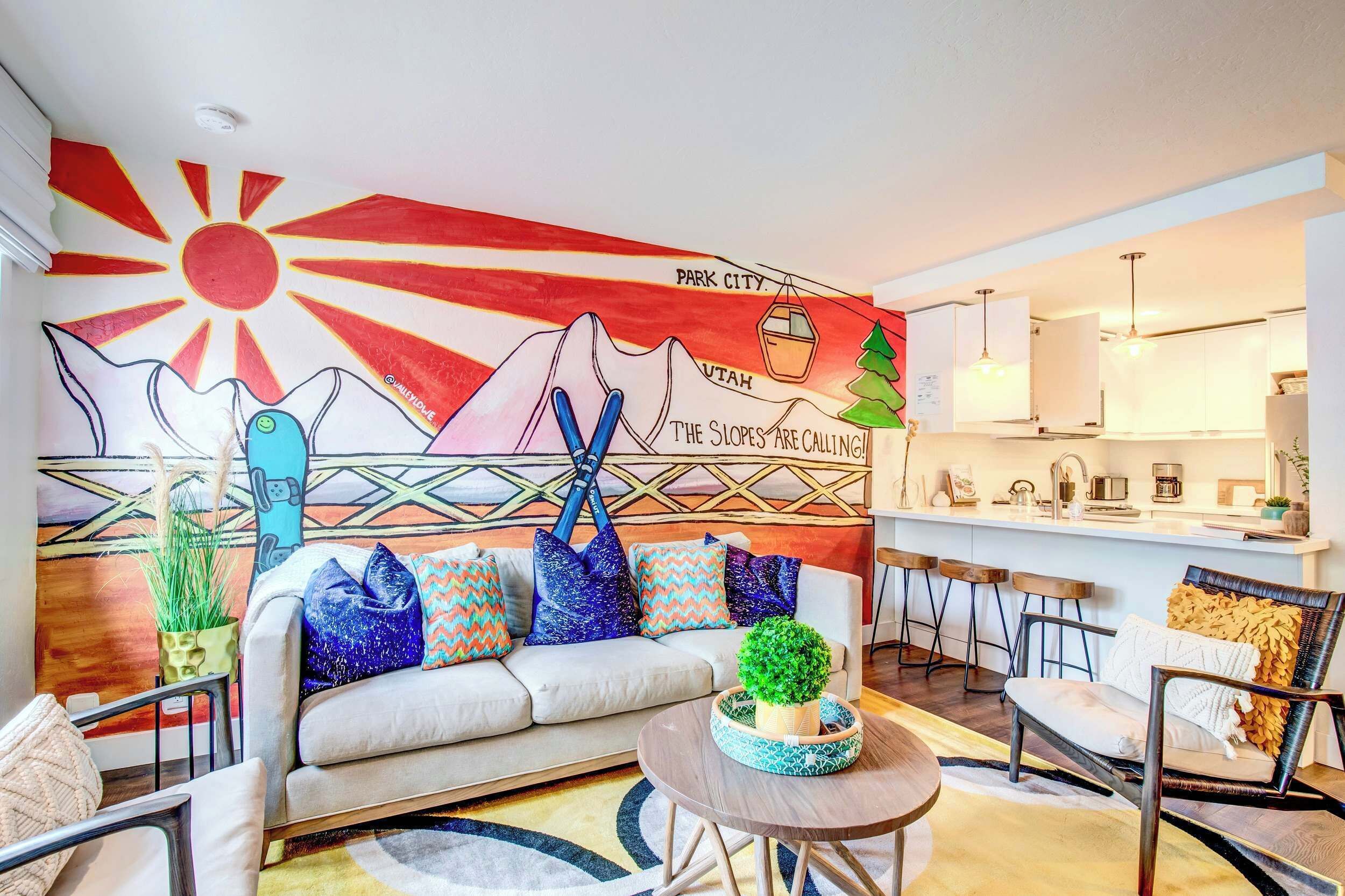Una sala de estar moderna con un mural colorido, un sofá, una mesa de centro y una cocina al fondo.