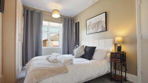Un letto rifatto con lenzuola bianche e una coperta beige, un comodino con una lampada, e un quadro con una veduta cittadina appeso al muro.