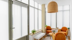 Een moderne kamer met grote ramen, een geweven hanglamp, een bankje en oranje stoelen.