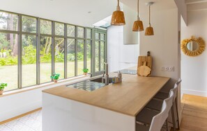 A modern kitchen with large windows, a wooden countertop, and hanging pendant lights. - Villa d'exception bord de mer (Noirmoutier en l'île)