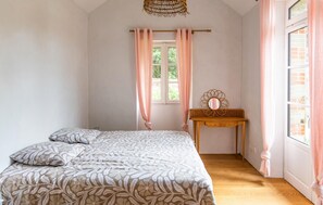A bedroom with a bed, a wooden side table with a mirror, and a window with curtains. - Villa d'exception bord de mer (Noirmoutier en l'île)