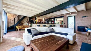 Un área de estar amplia con un sofá blanco, una mesa de centro de madera y un piso con baldosas de piedra. Se ve una escalera al fondo que conduce a un nivel superior.