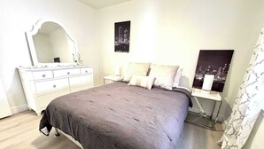 A bedroom with a large bed, a dresser, a nightstand, and a mirror. There are two wall-mounted pictures and a window with curtains. - Humble Haven - 2BR/1BA - Close to Hospitals (Lubbock)