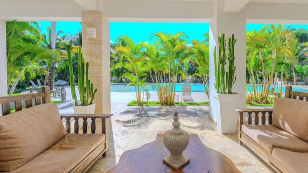 A poolside area with two sofas, a wooden table, and a view of a pool and tropical vegetation. - Residencial Paraiso Bayahibe (Bayahíbe)