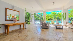 A spacious indoor area with a stone floor, a wooden bench, a sofa, a table, and a large mirror. There are potted plants and a view of a pool and palm trees outside. - Residencial Paraiso Bayahibe (Bayahíbe)