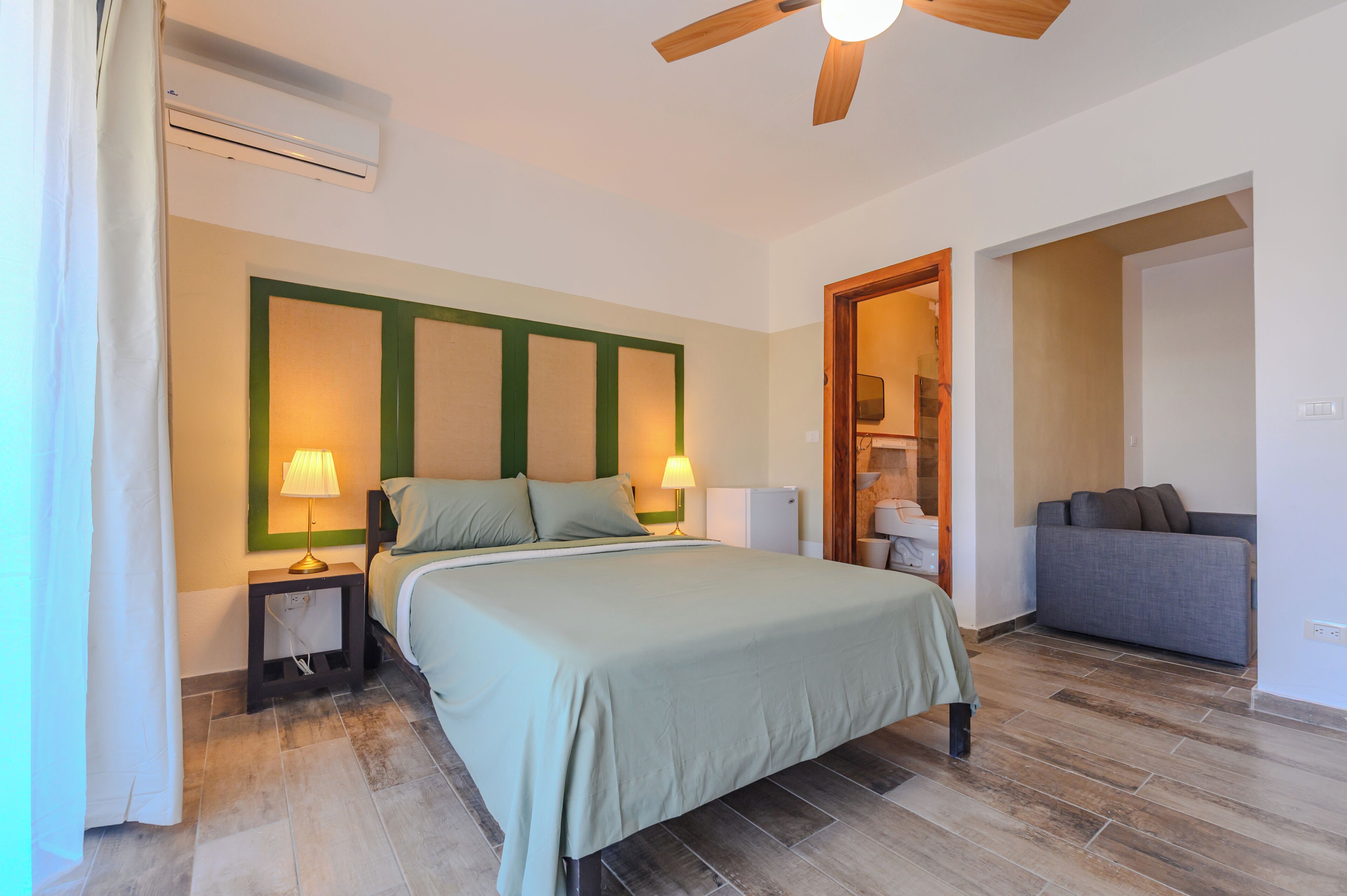 A bedroom with a bed, bedside lamps, a ceiling fan, and a sofa in the adjacent room.