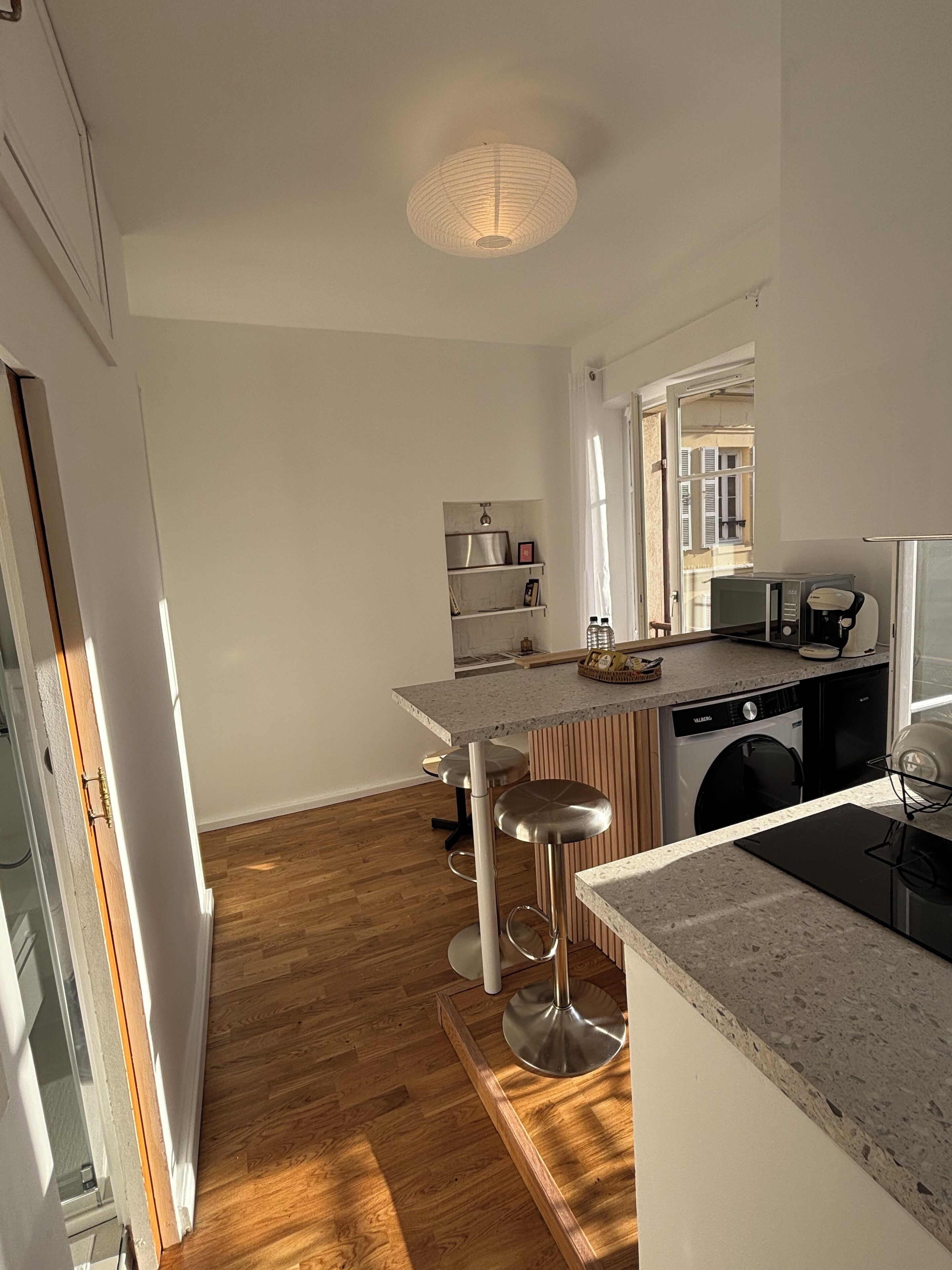 A modern kitchen with a central island, bar stools, a washing machine, and a dining area with a view of the outside.