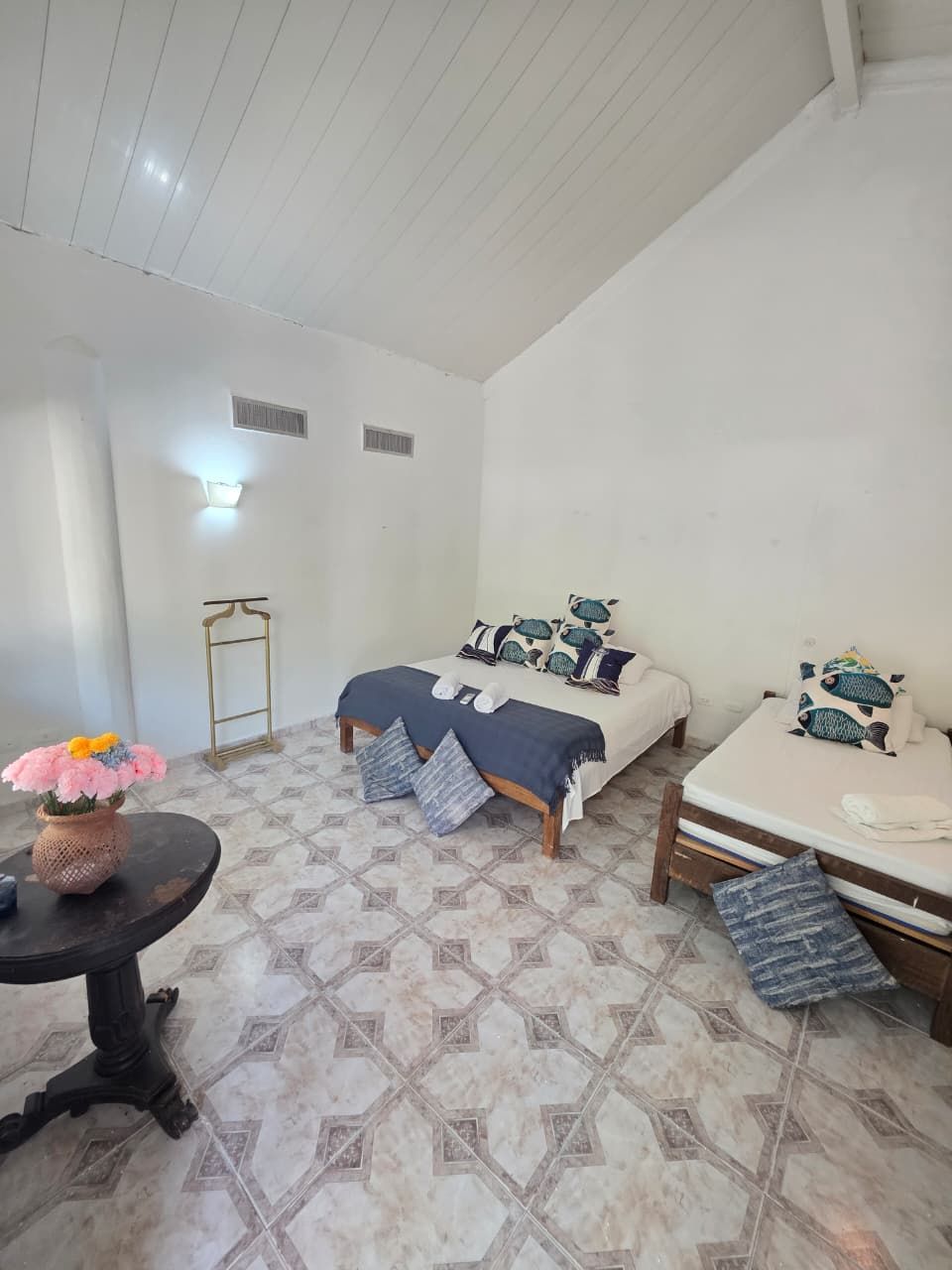 A room with two beds, a small table with a vase of flowers, and patterned floor tiles.