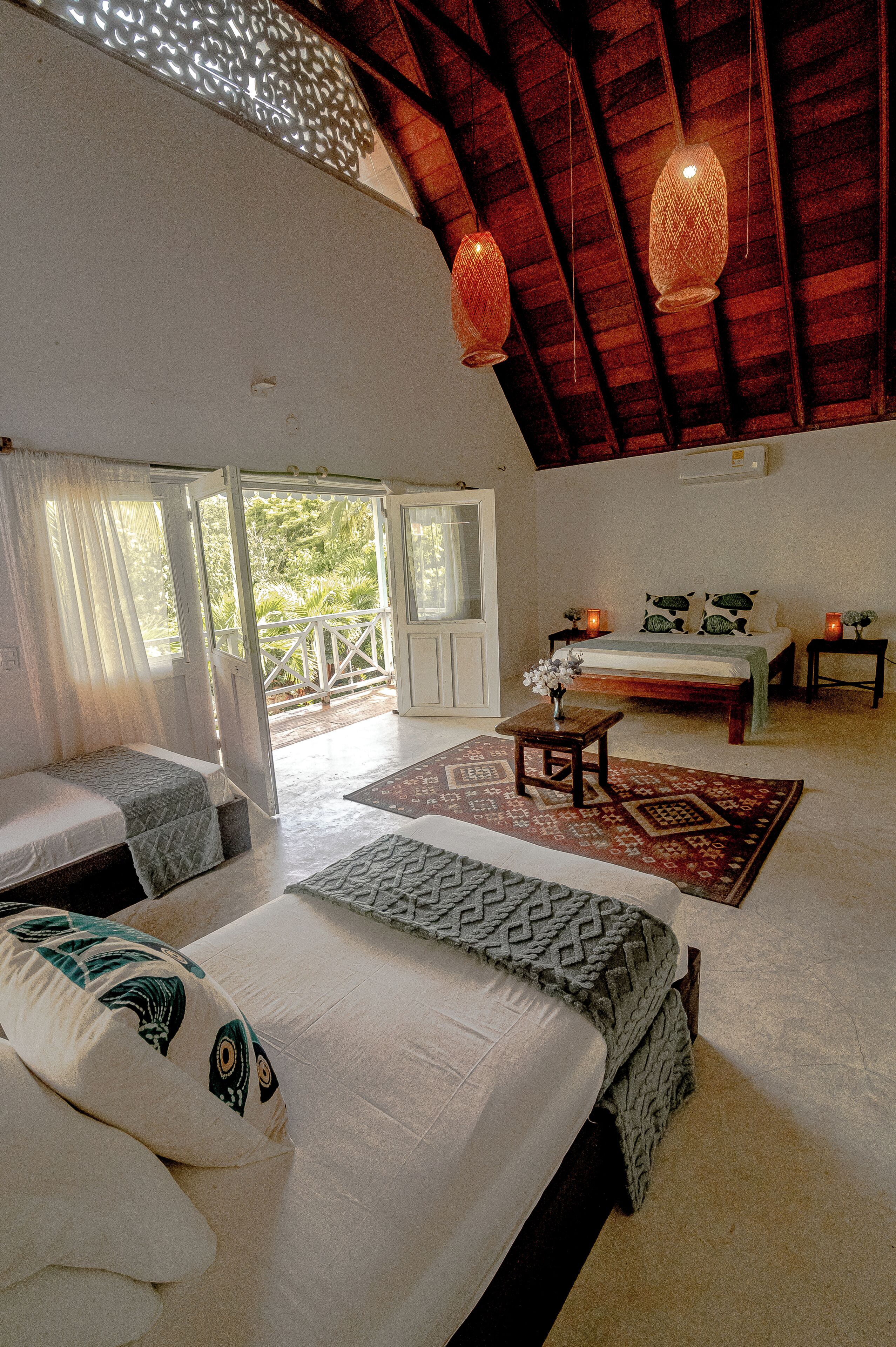 A room with a bed, a small table with a vase, a patterned rug, and a balcony with a railing.