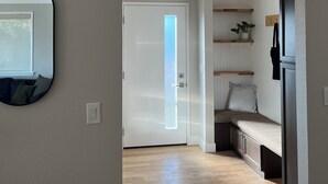 Un pasillo con piso de madera, una puerta blanca y un espejo a la izquierda. Hay estantes de madera con plantas y un banco con un cojín.