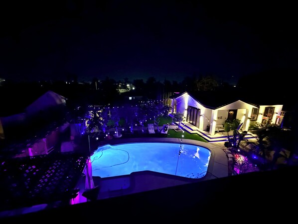 A swimming pool with a surrounding deck, a small garden, and a house with illuminated exterior lighting. - Heated Pool + EV Charger • Near Disney & Great Wolf  Central OC (Santa Ana)