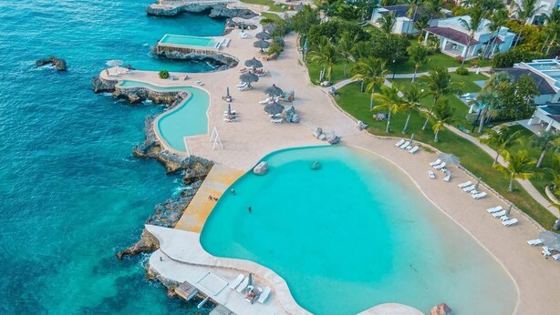An aerial view of a resort with a large turquoise pool, a sandy beach, and a rocky coastline. - Vive Tracadero Lodge - 2 Hab Apto (Dominicus)
