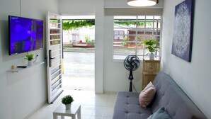 Una sala de estar con un sofá gris, un televisor montado en la pared, un ventilador y una ventana con vistas al exterior.