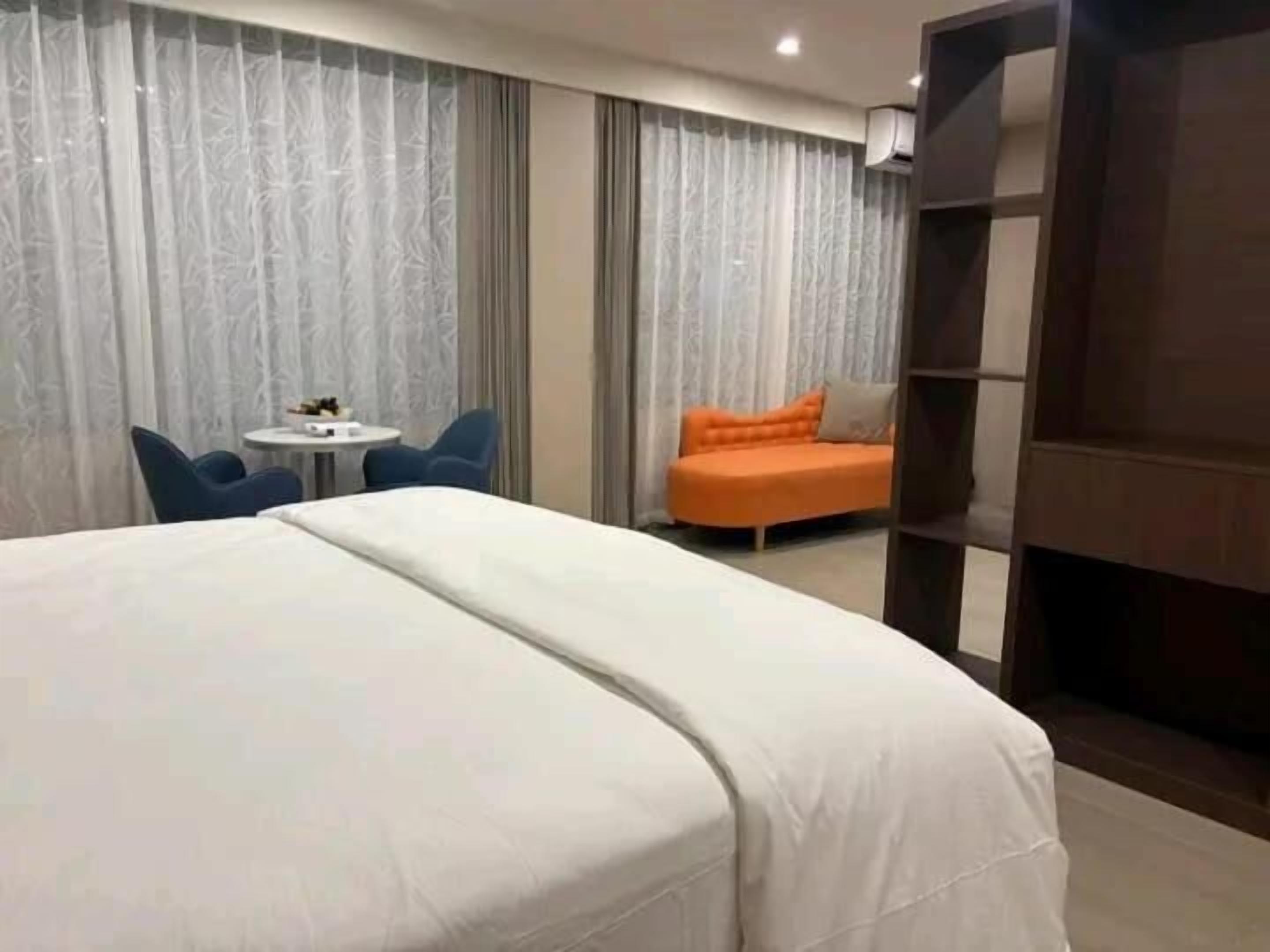 A hotel room with a large bed, a small round table, two chairs, an orange sofa, and a wooden bookshelf.