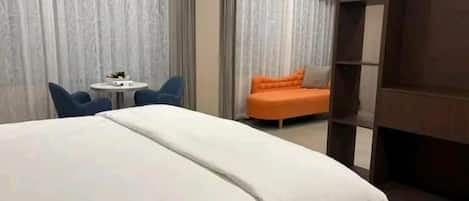 A hotel room with a large bed, a small round table, two chairs, an orange sofa, and a wooden bookshelf.