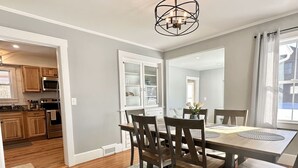 A dining area with a wooden table and chairs, a kitchen in the background, and a chandelier. - 3 BR Home in Ellsworth [High St Haven] (Ellsworth)