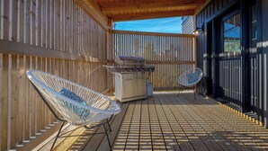 Une terrasse en bois avec un barbecue, deux chaises blanches et un espace abrité offrant une vue sur l’extérieur.