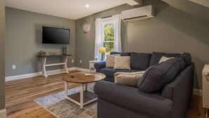 En stue med en mørkegrå sofa, et træ sofabord, et fladskærms-tv monteret på væggen og et vindue med hvide gardiner.