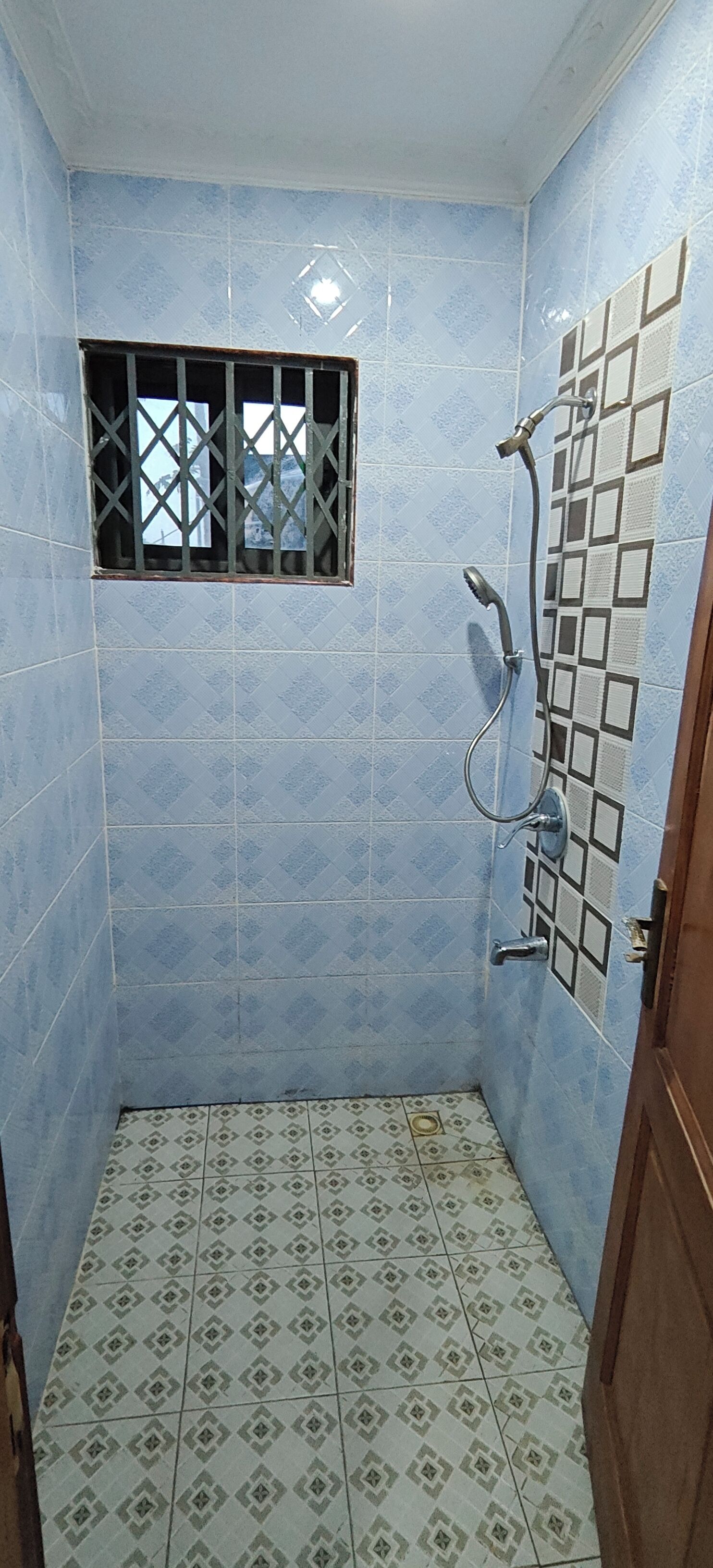 A small, tiled bathroom with a shower, a window with bars, and a patterned floor.