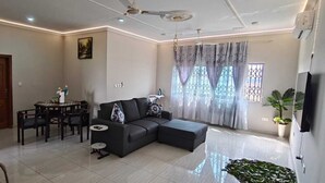 A living room with a sectional sofa, a dining table with chairs, a ceiling fan, and a large window with curtains.
