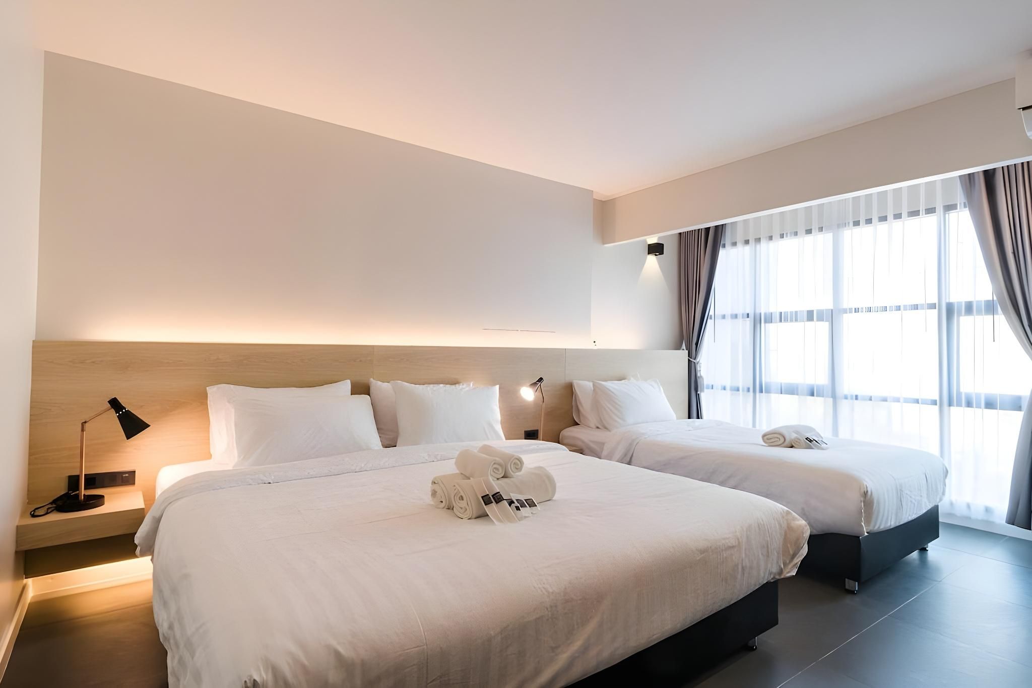A hotel room with two beds, a bedside table with a lamp, and a large window with curtains.