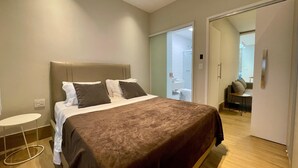 A modern bedroom with a large bed, a bedside table, and a view into the bathroom and living area. - Beachfront apartment, two suites, Post 9 - Ipanema (Rio de Janeiro)