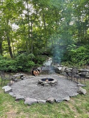 A circular stone fire pit surrounded by a gravel area, with a metal grill and chairs.