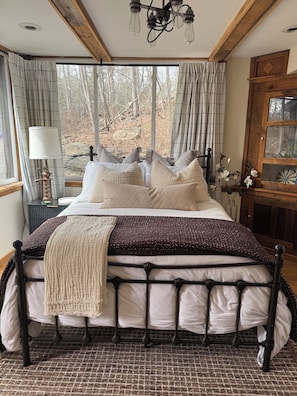 A bed with a metal frame, a woven blanket, and a view of trees through a window.