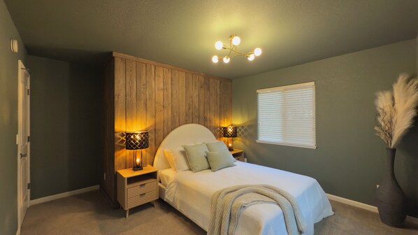 A bedroom with a bed, two bedside tables, a wooden wardrobe, a window with blinds, and a ceiling light fixture.