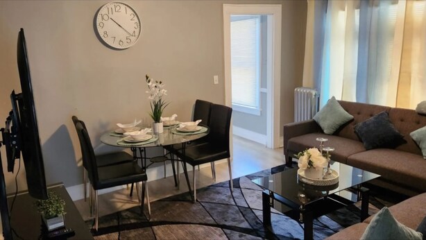 Ein Wohnzimmer mit Sofa, Couchtisch, Esstisch mit Stühlen, einer Wanduhr und einem Fenster mit Jalousien.