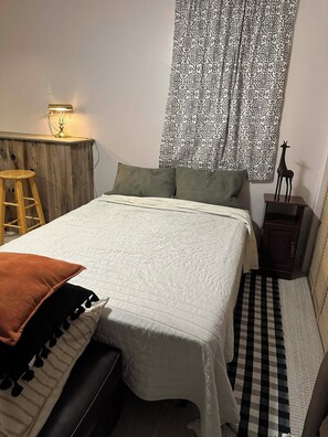 Une chambre à coucher avec un lit, une table de chevet en bois, une lampe, un petit tabouret, un rideau à motifs et une figurine décorative représentant une girafe.