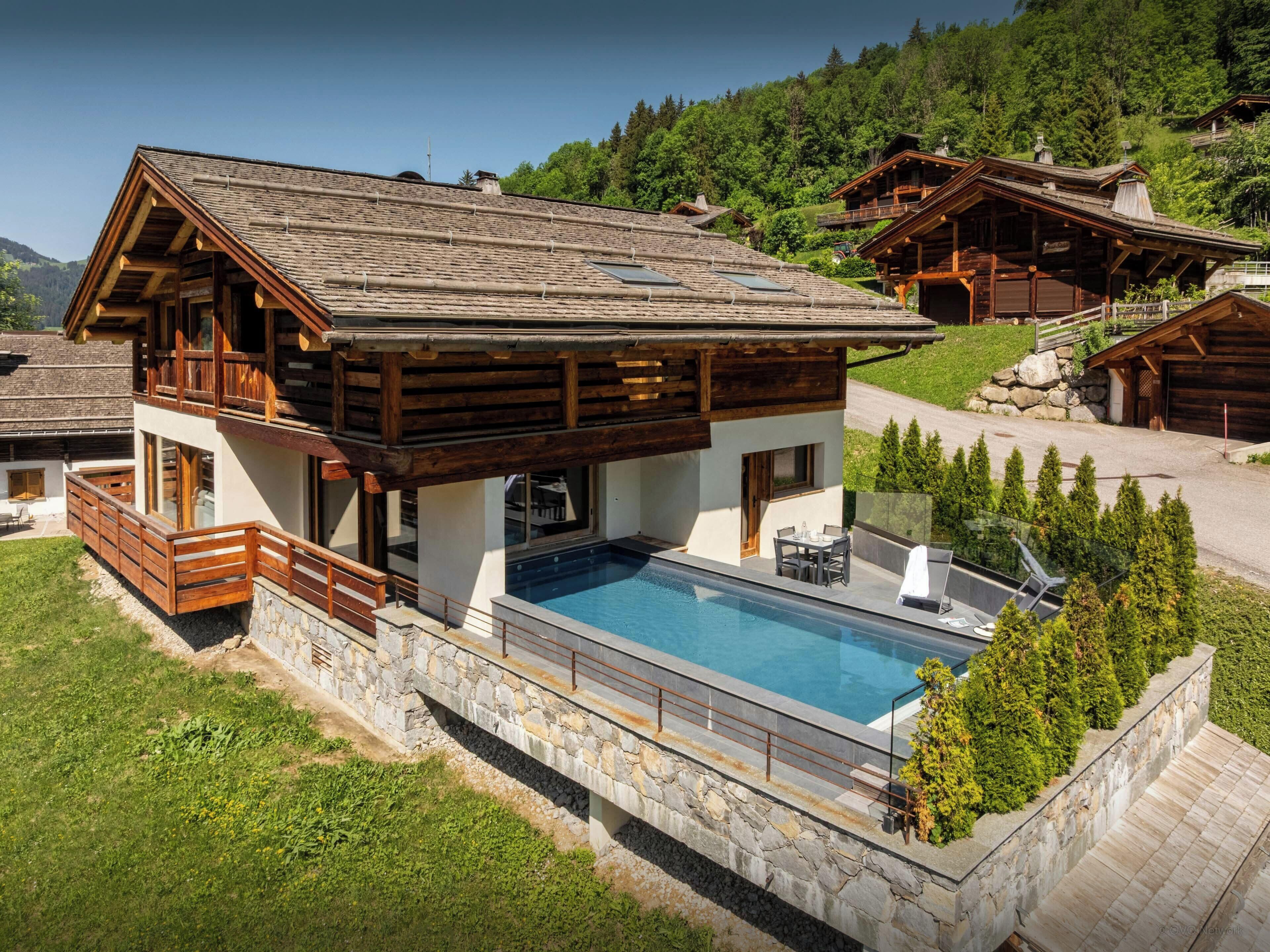 A wooden chalet with a swimming pool, stone walls, and a view of the surrounding forest.