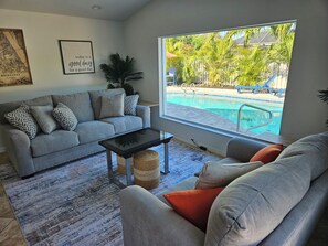 A living room with a grey sofa, a coffee table, and a view of a swimming pool outside. - Peaceful Beachside, Family Home w/ Heated Pool! (Satellite Beach)