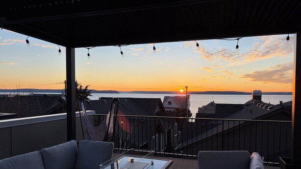 Ein Balkon mit Glastisch und zwei Sofas, mit Blick auf einen Sonnenuntergang über dem Wasser und Gebäuden im Hintergrund.