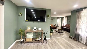 Una sala de estar con un televisor de pantalla plana grande montado en la pared, una mesa consola de madera con adornos, un sofá de cuero marrón y una alfombra estampada en el suelo.