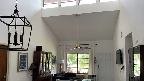 Een ruime woonkamer met een hoge plafond, een kroonluchter, een plafondventilator, een televisie en een groot raam met gordijnen.