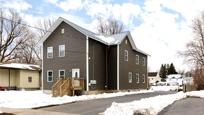 Un edificio residencial de dos pisos con revestimiento exterior oscuro, un porche con escalera de madera y un camino de entrada cubierto de nieve.