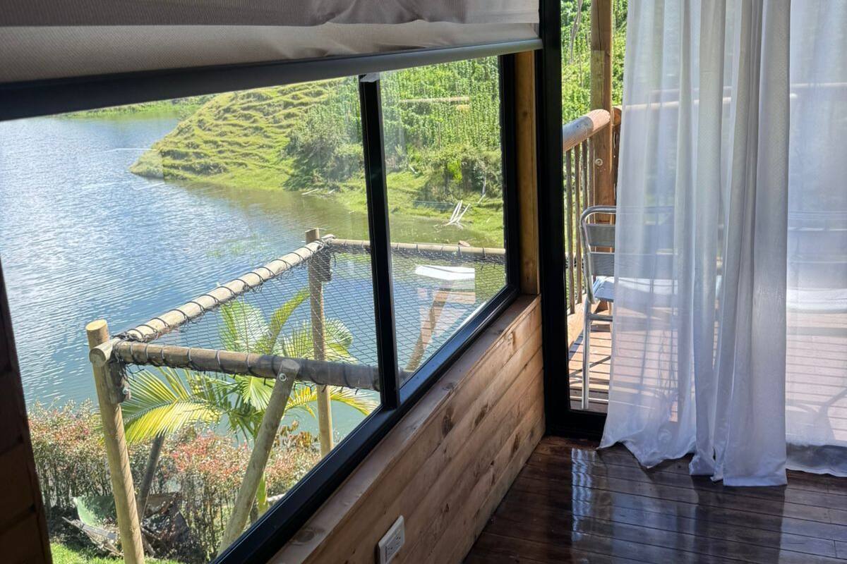 A room with a large window overlooking a water body and greenery, wooden walls, and a wooden deck outside.