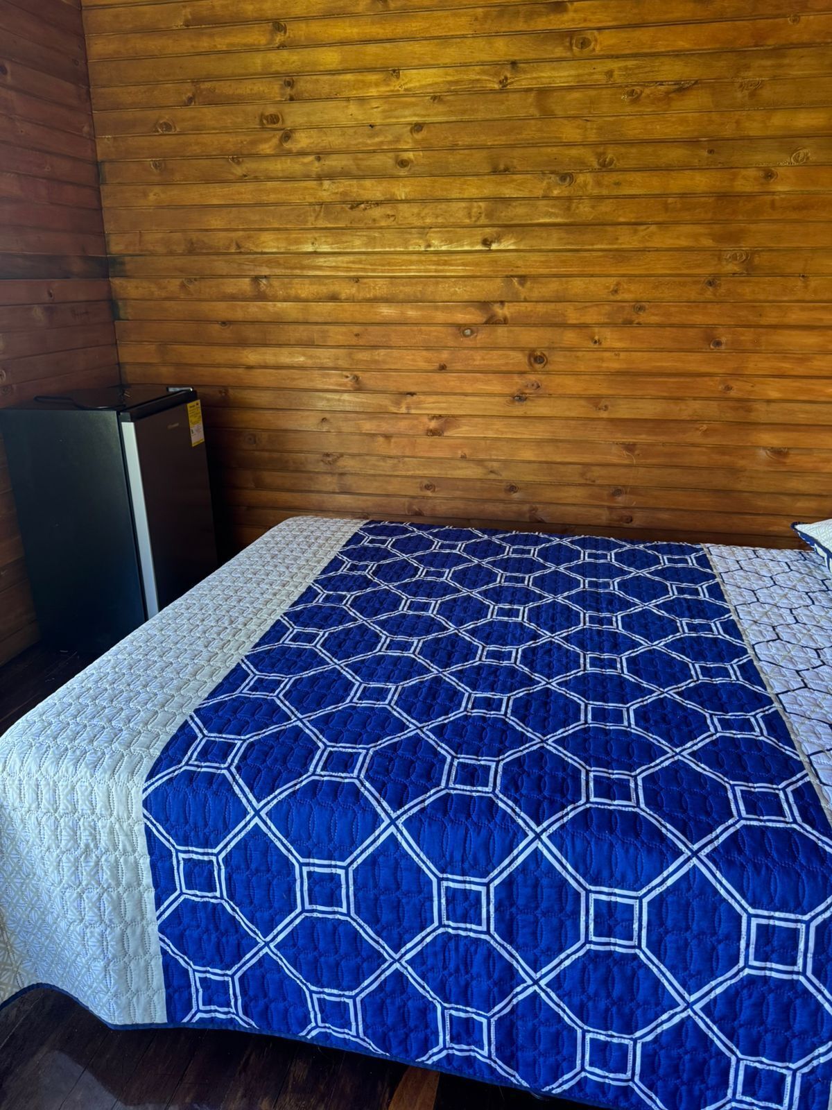 A bed with a blue and white quilt, a wooden wall, and a small black refrigerator.