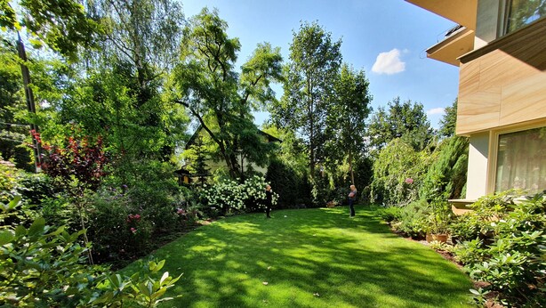 Ein gepflegter Garten mit vielfältigen Pflanzen und Bäumen, einem ordentlich gestutzten Rasen und einem modernen Gebäude mit großen Fenstern.