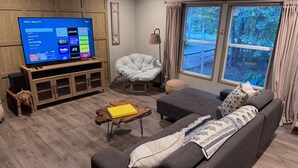 A living room with a large flat-screen TV, a grey sofa, a wooden coffee table, and a chair.