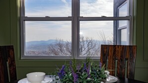 Ein Esstisch mit einer weißen Schüssel und Untertasse, einem Topf mit violetten Blüten und Blick auf Berge durch ein Fenster.