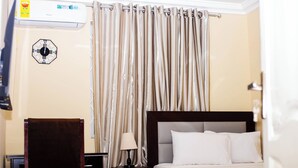 A hotel room with a bed, a chair, a nightstand with a lamp, an air conditioning unit, and a curtain. - De GraceManuel Terrace (Gbawe)