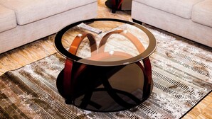A round coffee table with a glass top and wooden legs, placed on a patterned rug in a living room with a beige sofa and a small round table in the background. - De GraceManuel Terrace (Gbawe)