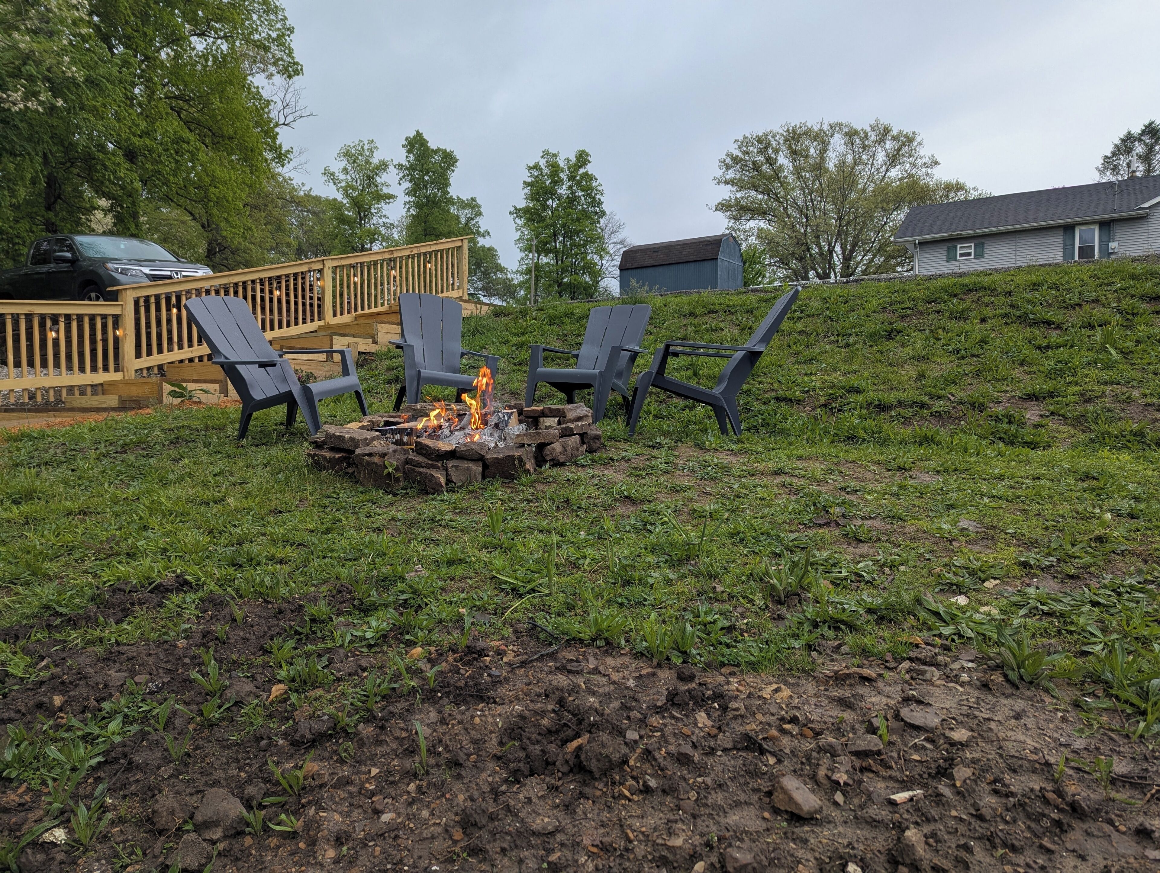 Ein Garten mit Feuerstelle, umgeben von vier Stühlen, einer Holzterrasse und einem Haus im Hintergrund.