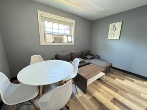 Une pièce avec une table ronde, des chaises blanches, un canapé gris, un banc en bois, un radiateur et une fenêtre avec des stores.