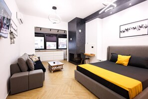 A modern bedroom with a large bed, a sofa, a small table, and a kitchen area visible through an open door. - Grandstay Central in Durr s (Durrës)