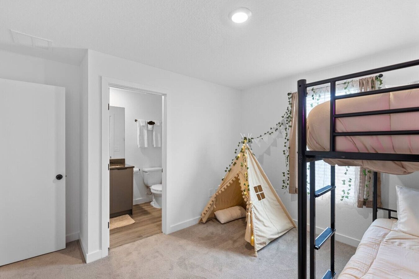 Un dormitorio con literas, un espacio de juego tipo tienda de campaña, un baño con inodoro y lavamanos, y una puerta que da a otra habitación.